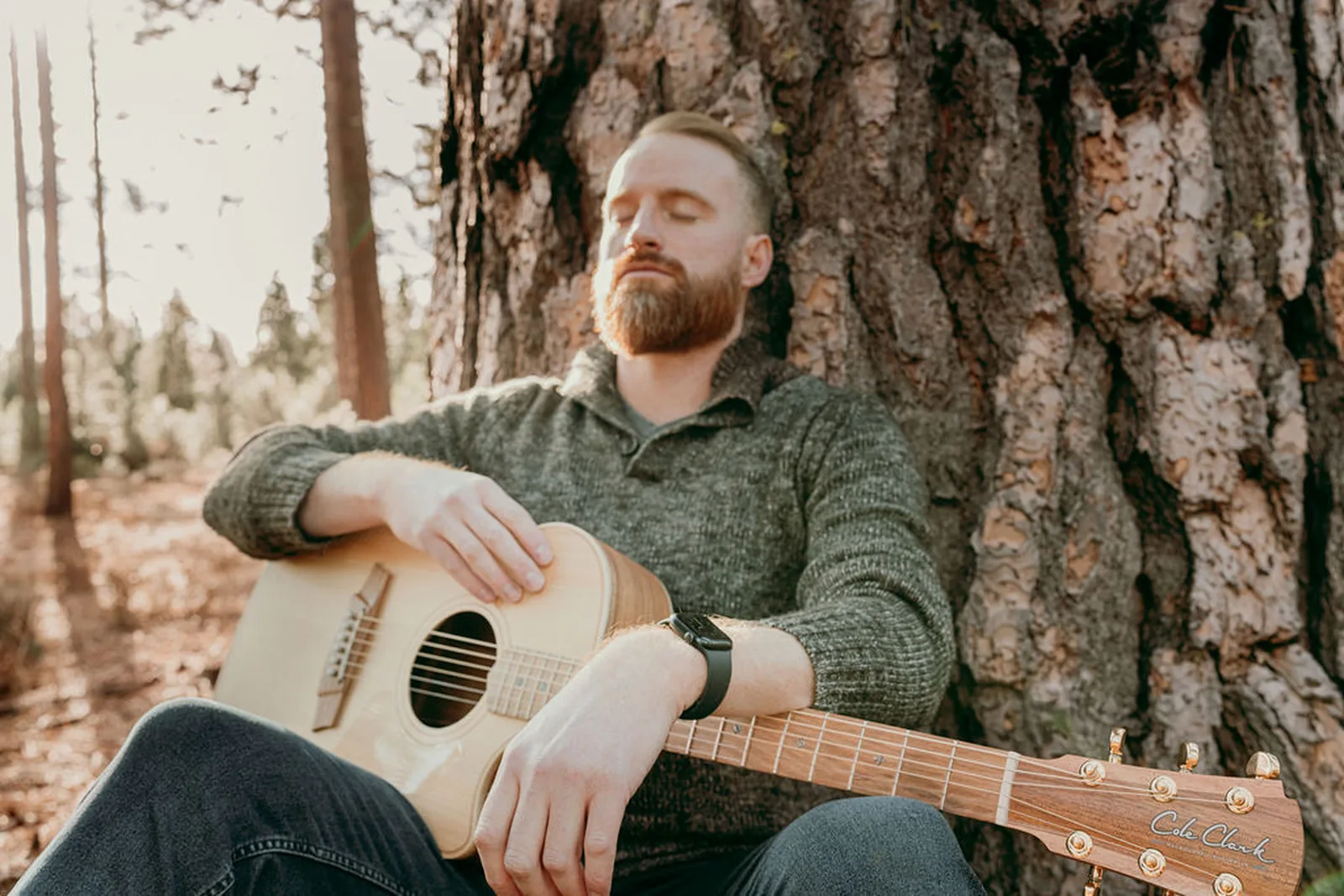 Roger seated against a pine with his guitar, eyes closed
